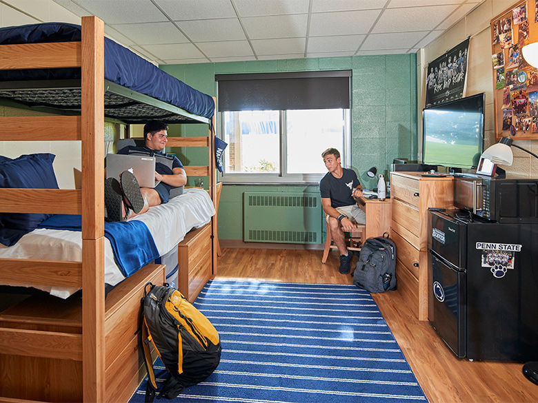 Two male students talk in a residence hall room. One is on a bunk bed, the other is at a desk.