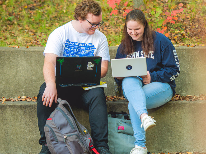 Two students dressed in Penn State t-shirts look at their computers while siting on steps outside.