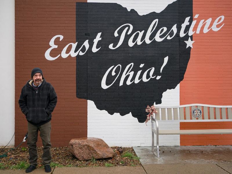 A man wearing a dark winter coat and a knit hat stands in front of a mural that says East Palestine, Ohio.
